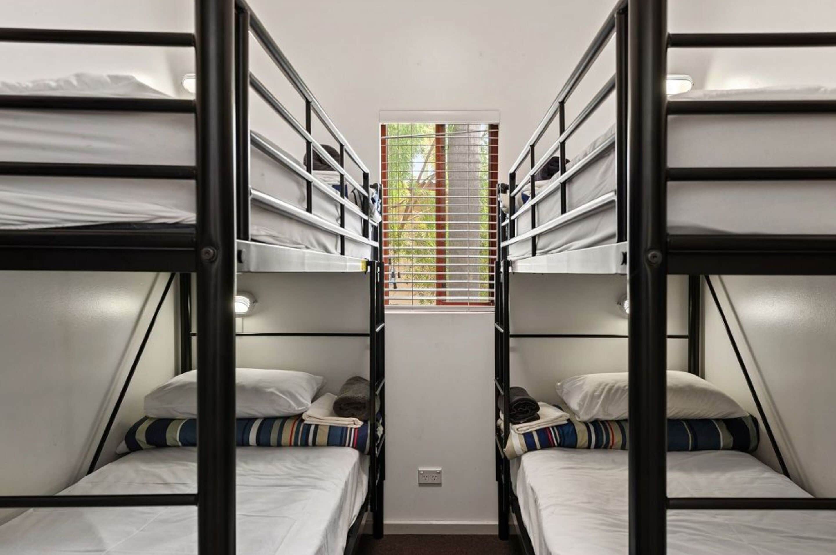 A bunk bed room with two beds, a window with blinds, and a small table in the center.
