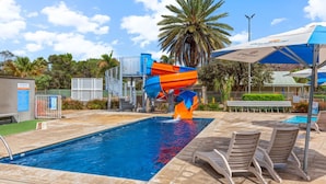 A swimming pool with a water slide, lounge chairs, and palm trees in the background. - Discovery Parks - Alice Springs (Ross)