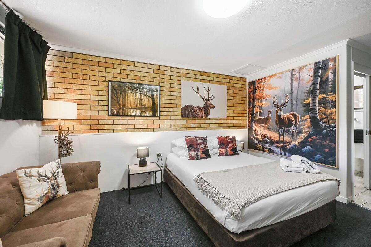 Une chambre avec un mur en briques, un lit recouvert d’une couette blanche, deux coussins à motifs de cerfs, une table de chevet avec une lampe et une grande fresque représentant une forêt sur le mur.