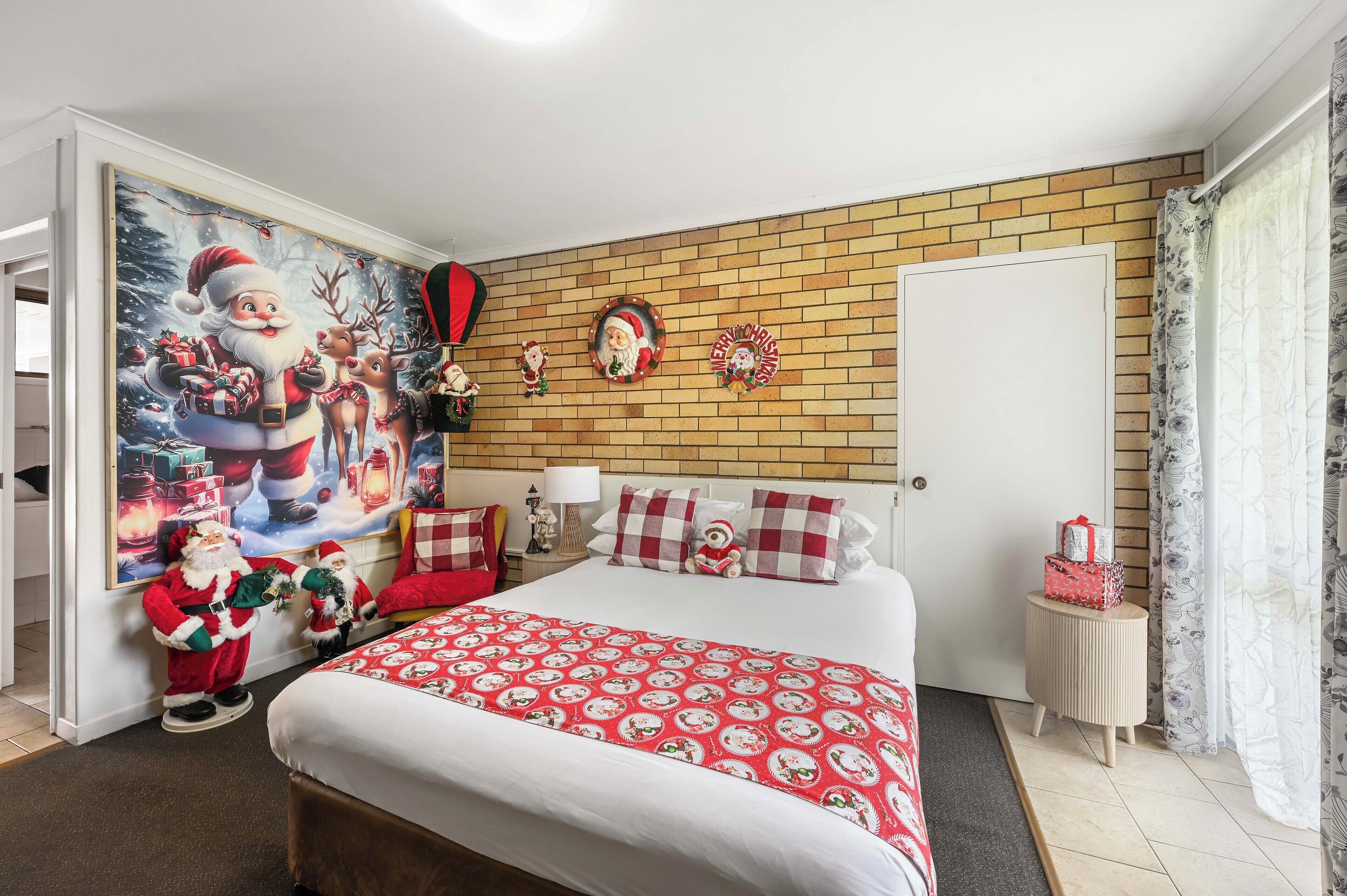 Un dormitorio con una cama grande, pared con diseño de ladrillos, una puerta, mesita de noche pequeña y decoración navideña en la pared.