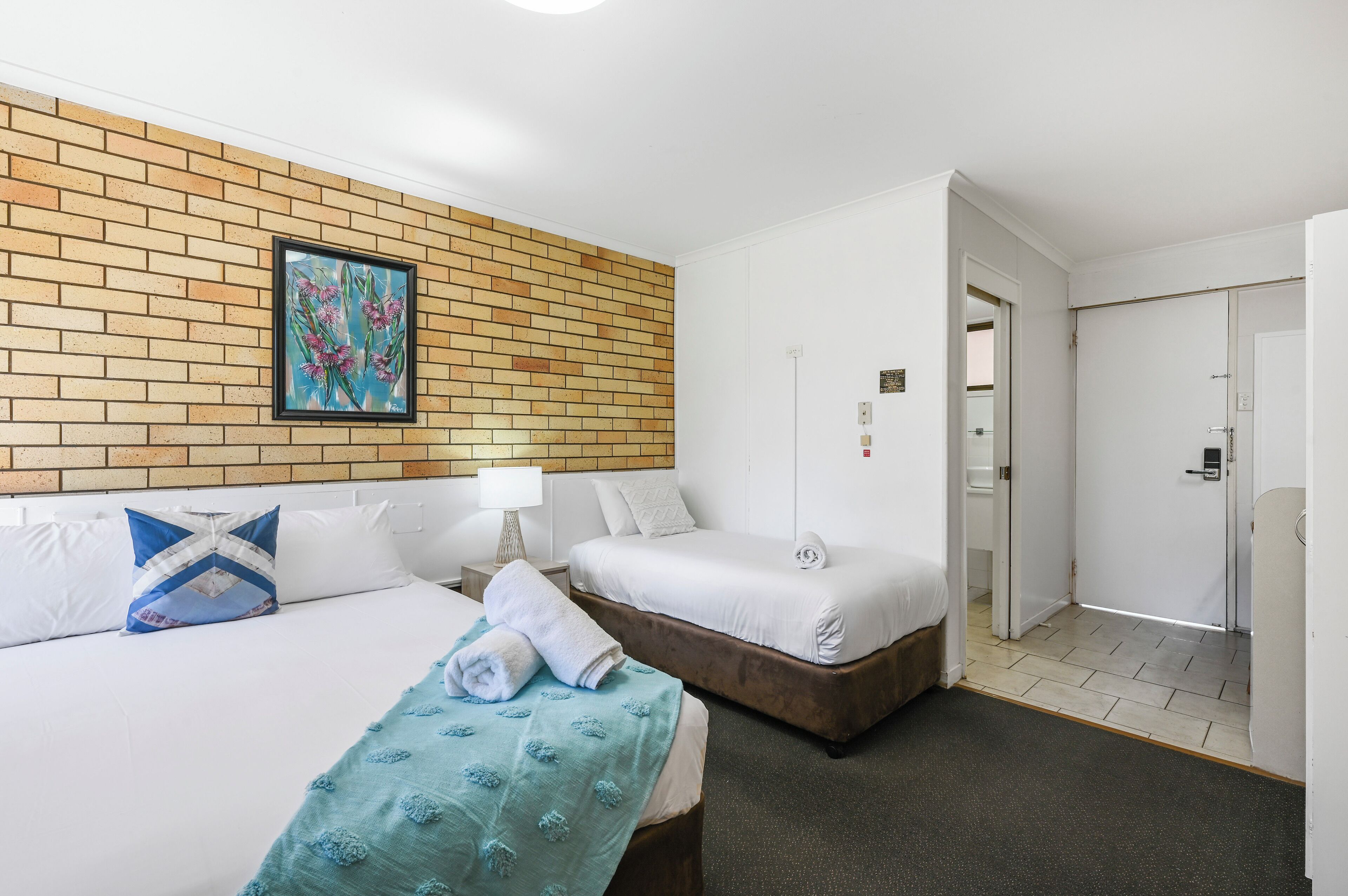 Habitación de hotel con dos camas, una pared de ladrillo decorativa, un cuadro y una puerta que da al baño.