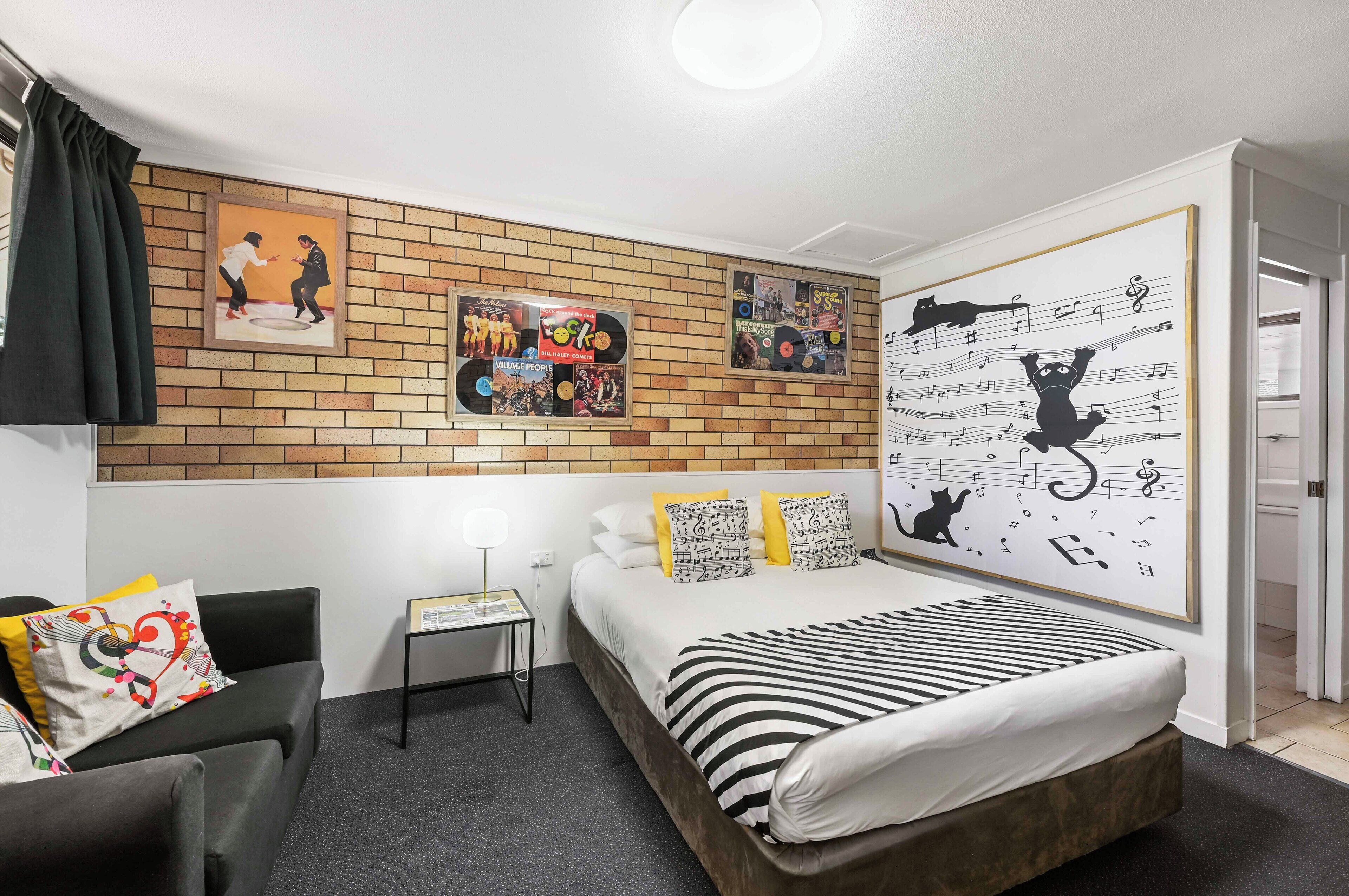 Una habitación de hotel moderna con una pared decorativa de ladrillo, una cama con ropa de cama a rayas, una mesita de noche pequeña y una silla con cojines de colores.