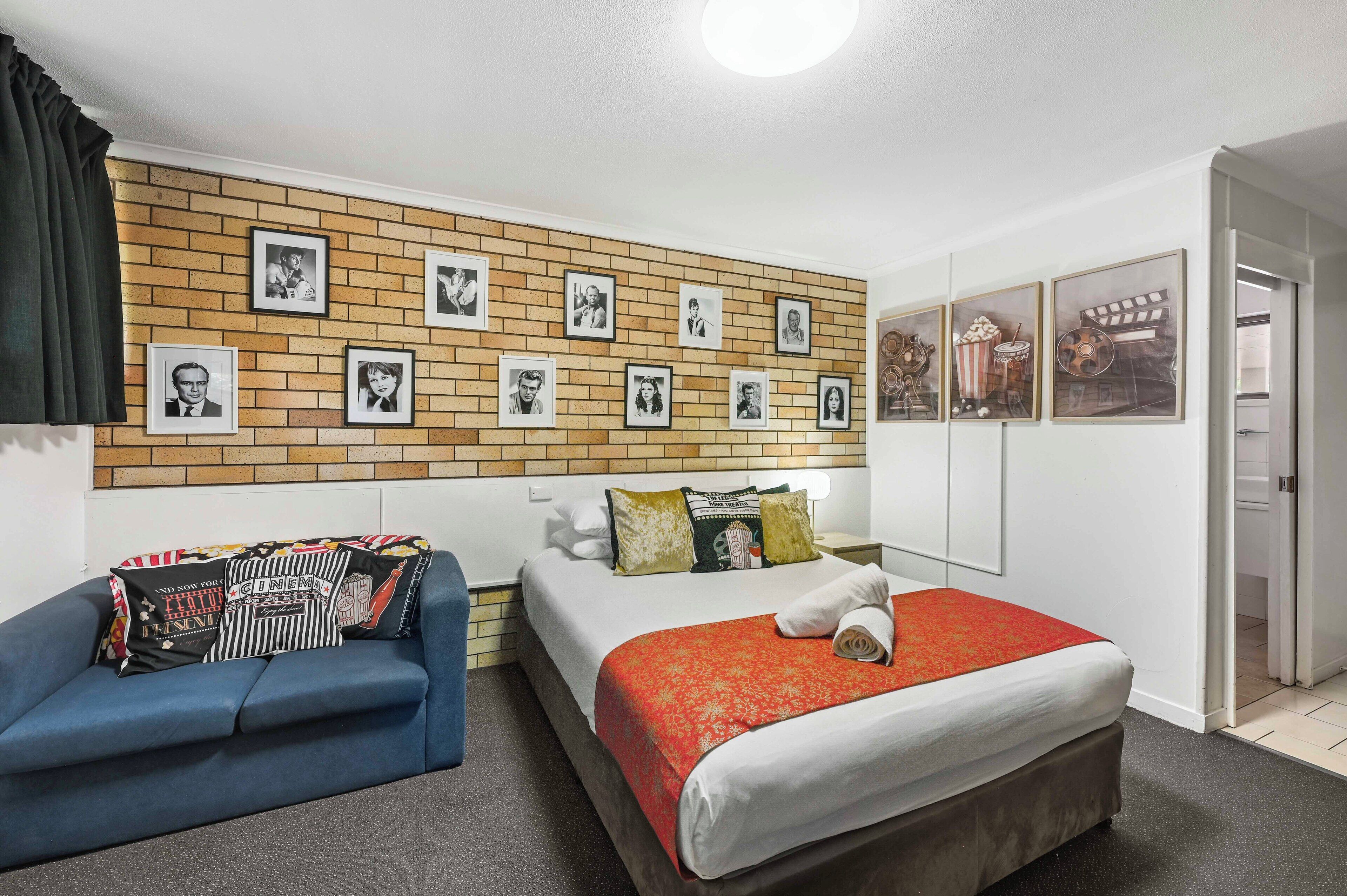 A hotel room with a brick-patterned wall, a bed with a red blanket, a blue sofa, and framed pictures on the wall.