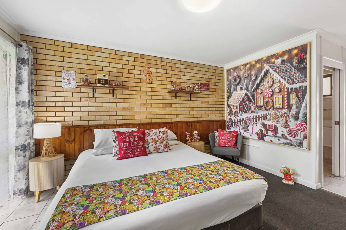 Une chambre d’hôtel avec un mur décoré d’un motif en briques, un grand lit avec une literie aux couleurs vives, une table de chevet avec une lampe et une fresque murale représentant une maison en pain d’épices.