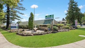 A motel with a green roof, a signboard, and a landscaped garden with a fountain.