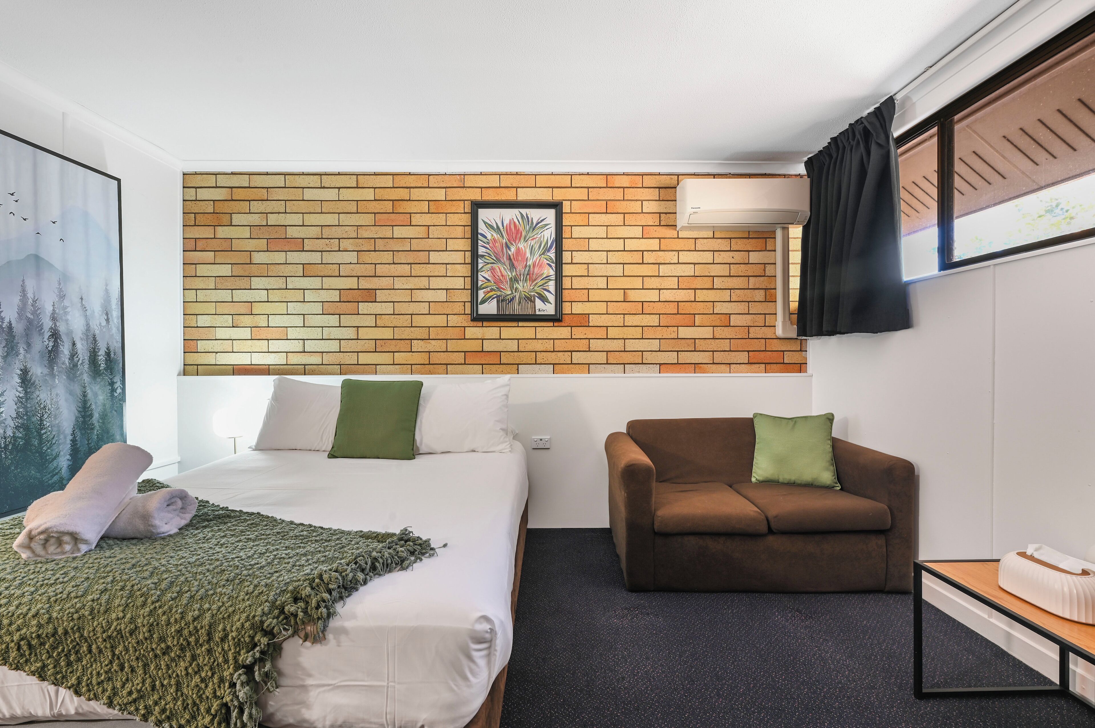 Habitación de hotel con cama, sofá marrón, una mesita pequeña y aire acondicionado instalado en la pared.