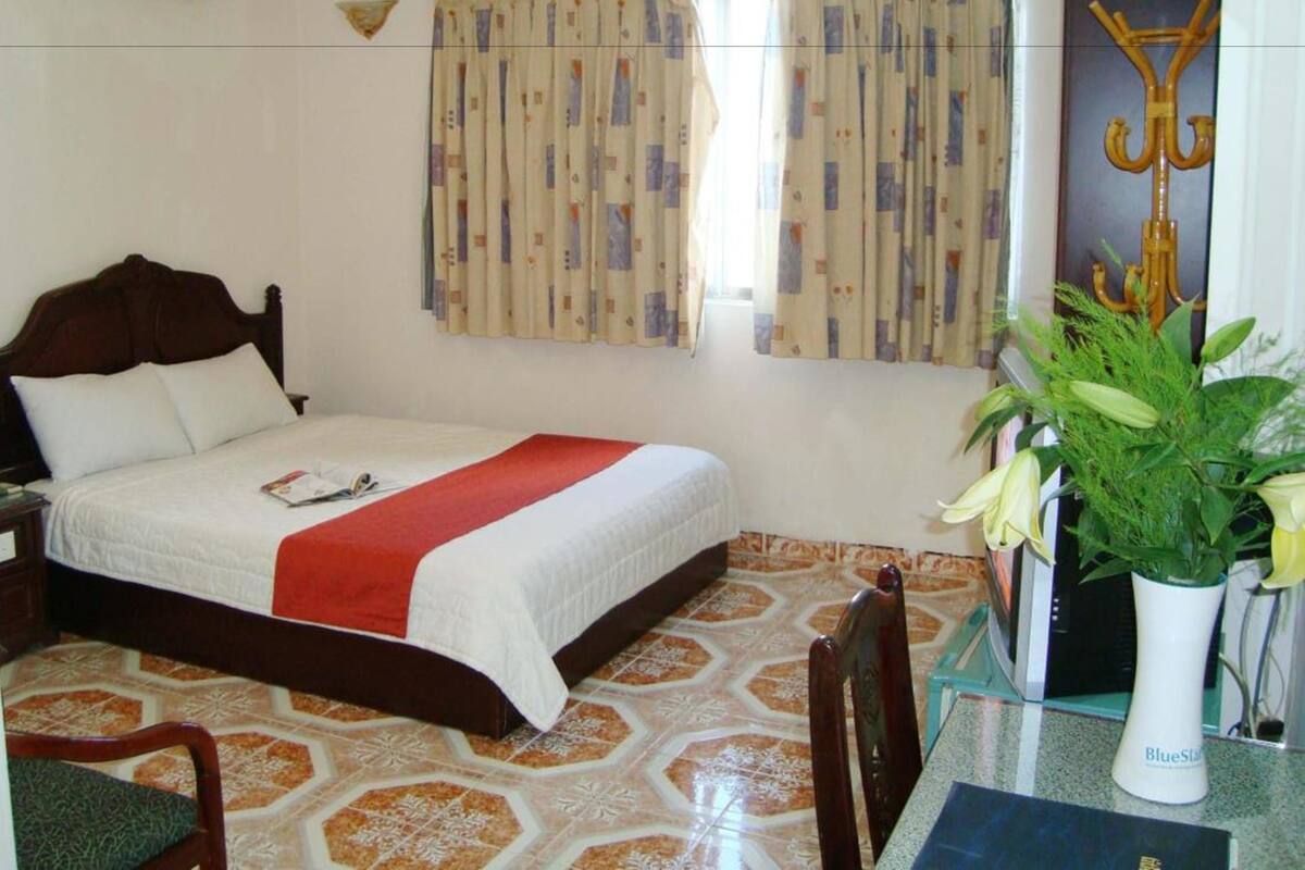 A hotel room with a bed, a chair, a table with a vase, and a window with curtains.
