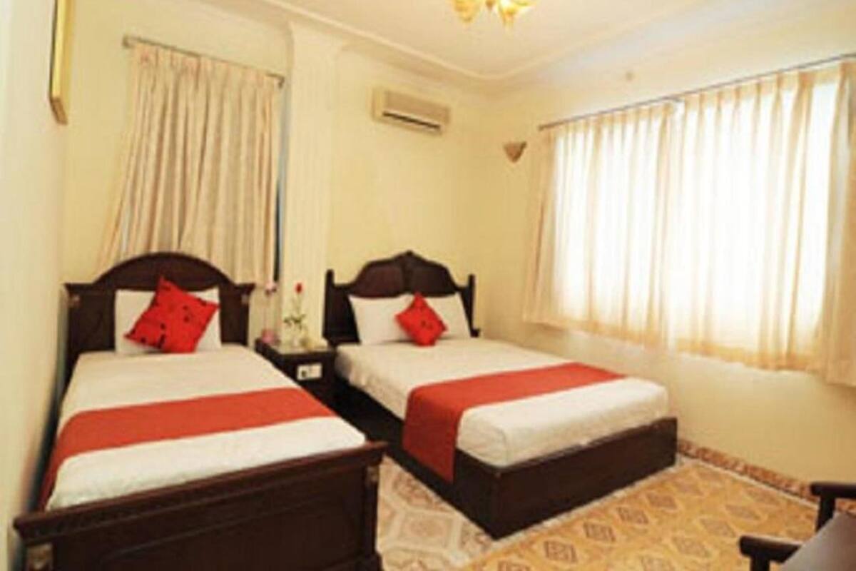A hotel room with two single beds, each with a red pillow and a white bedspread. There is a small bedside table with a lamp, a window with curtains, and a patterned carpet.