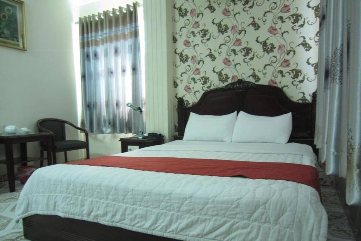 A neatly made bed with a white quilt and a red blanket, a wooden nightstand with a lamp, a chair, and a window with curtains.