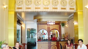 Un hall de hotel con varios relojes que muestran la hora de diferentes ciudades, un mostrador de recepción con un recepcionista y varios comensales sentados en mesas.