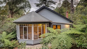 A house with a large glass bay window, surrounded by lush greenery and trees. - Garden Cottage at Fern Falls Cottages (Mount Dandenong)