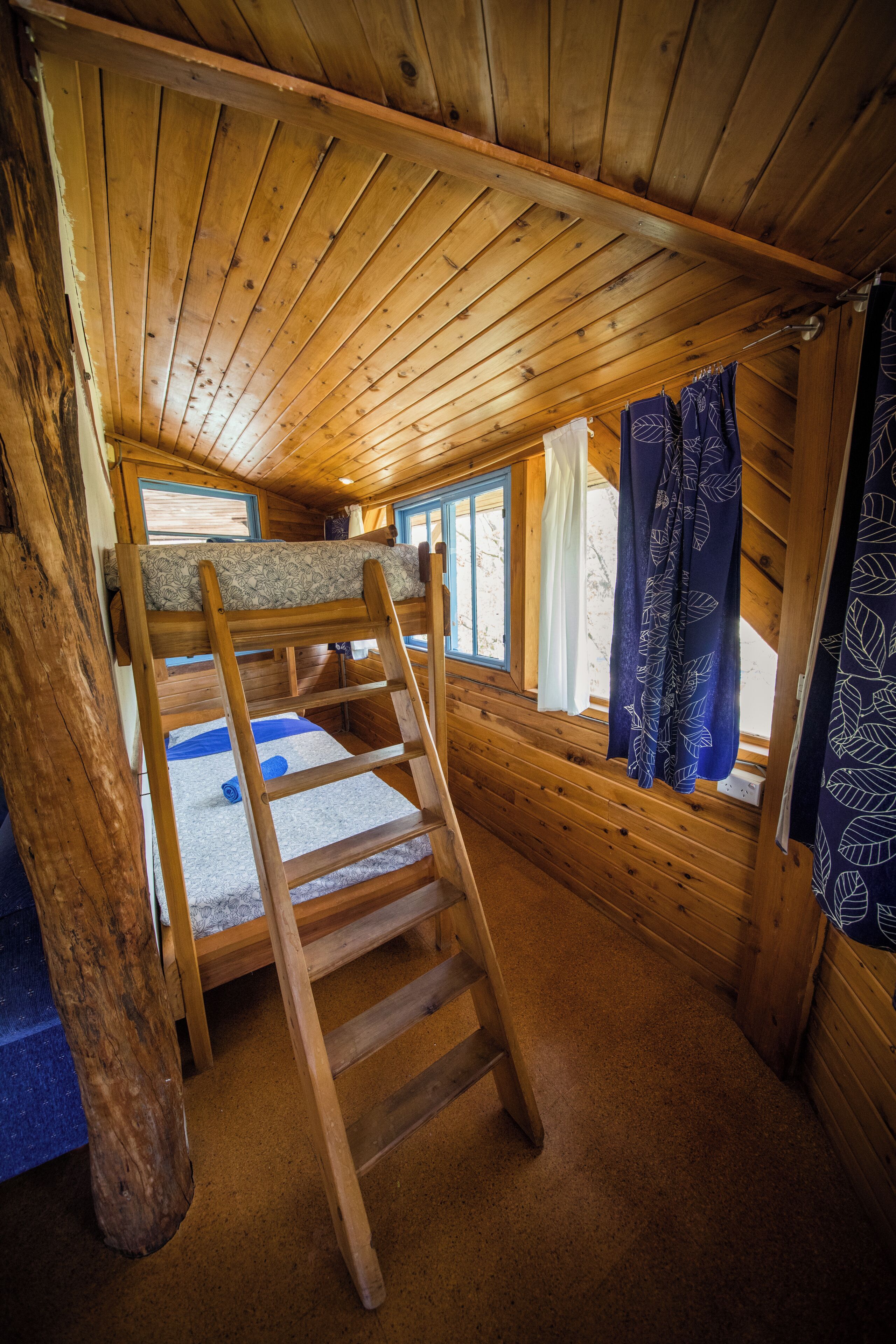 A wooden cabin room with a bunk bed, a ladder, a window with curtains, and a wooden ceiling.