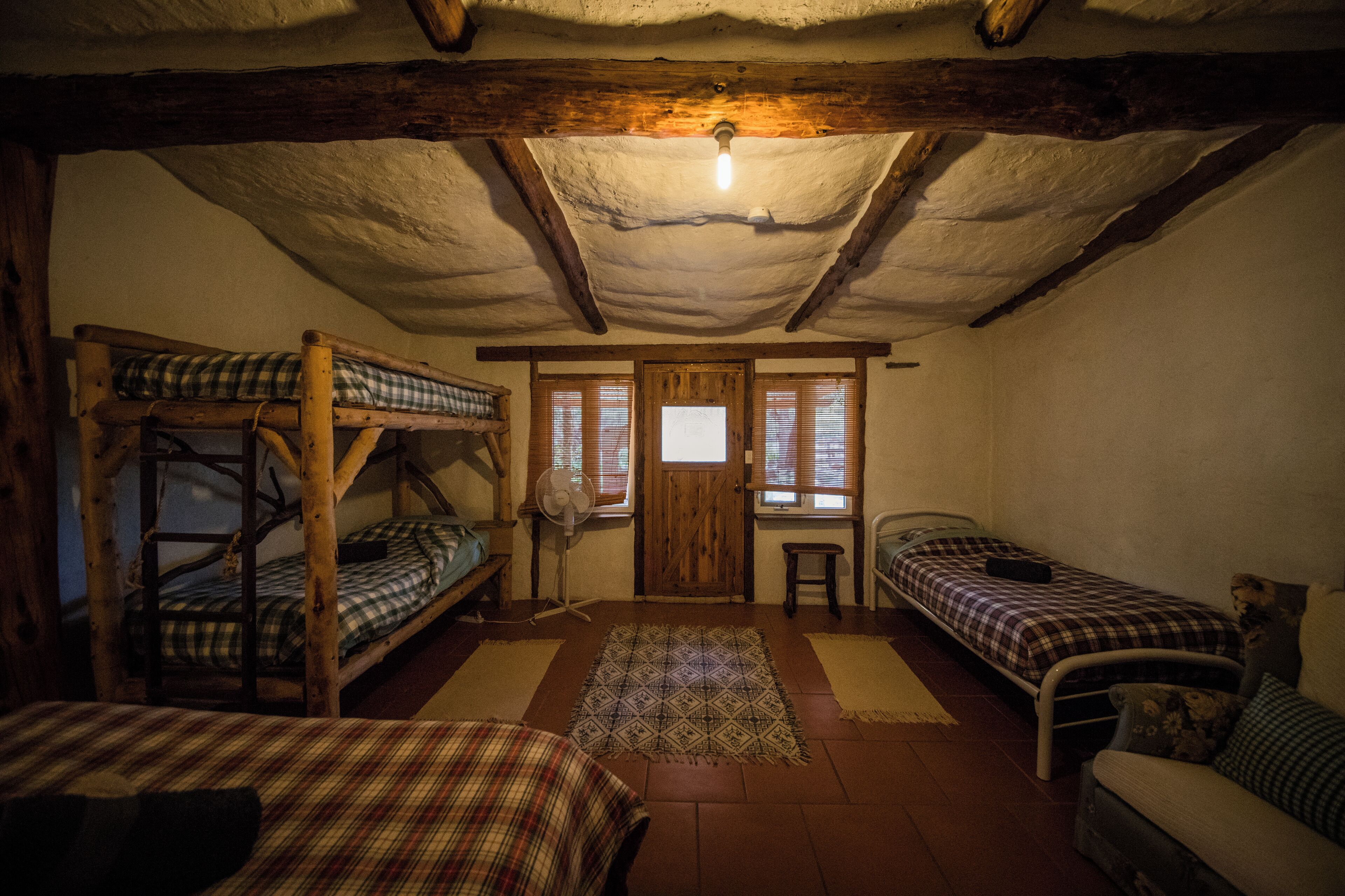 Una habitación rústica con dos literas y una cama individual, vigas de madera en el techo, una mesita y una puerta con ventanas.