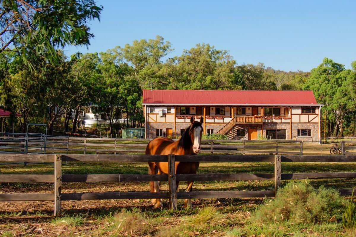 Un establo con techo rojo y paredes de madera, rodeado por una cerca con un caballo adentro.