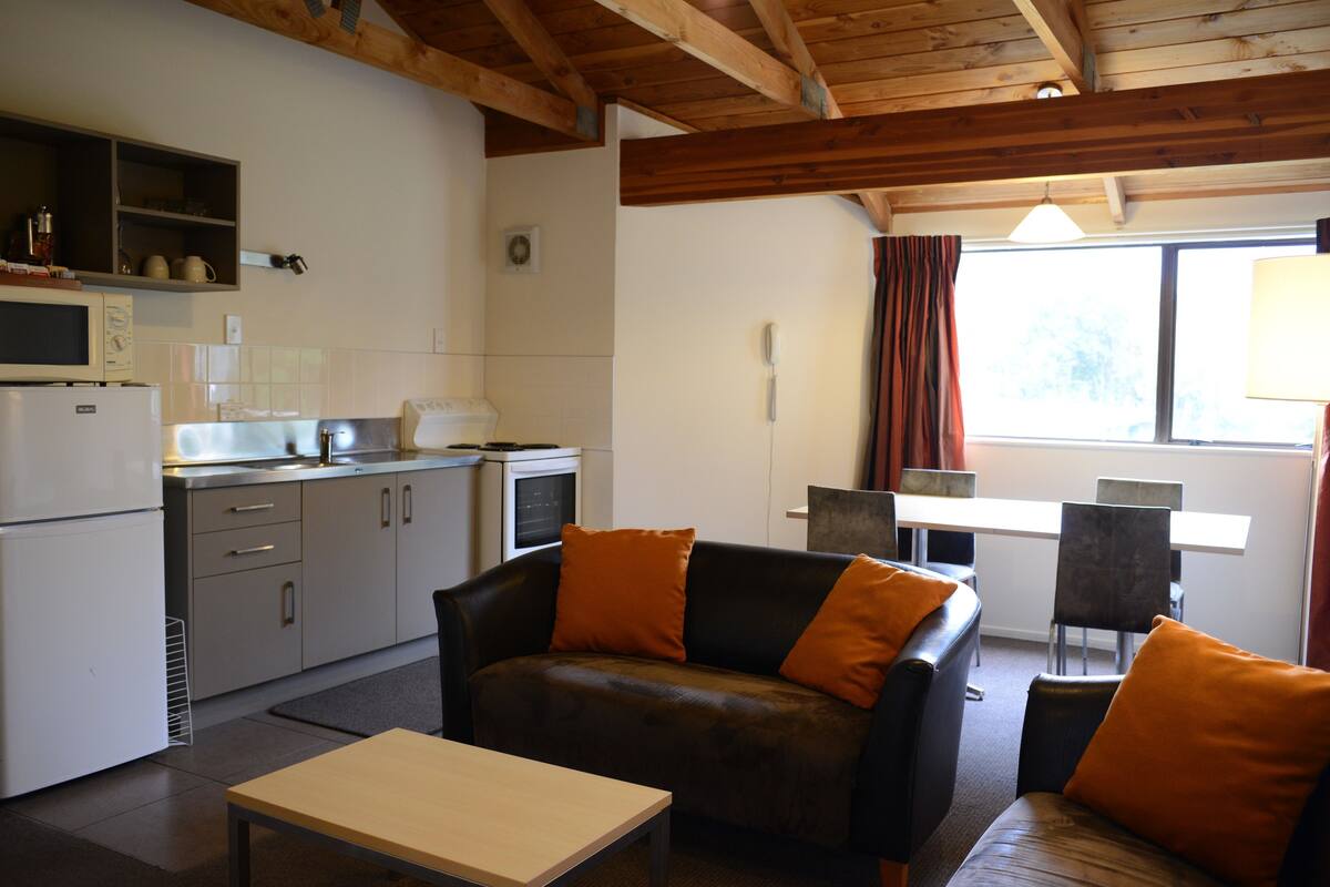 Cocina compacta con microondas, refrigerador y lavamanos. Hay un sofá con cojines naranja y una mesa de comedor con sillas. La habitación tiene vigas de madera en el techo y una ventana con cortinas.