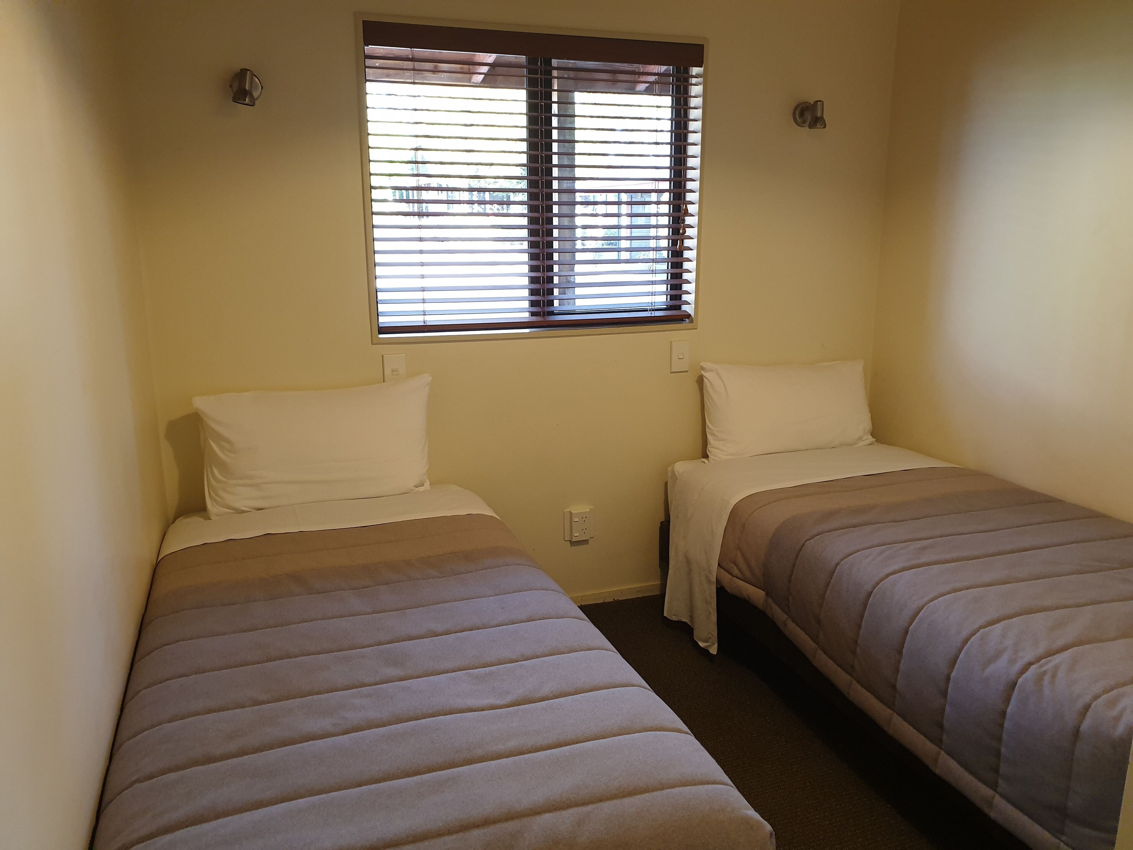 A small, single-room accommodation with two single beds, a window with blinds, and a plain wall.
