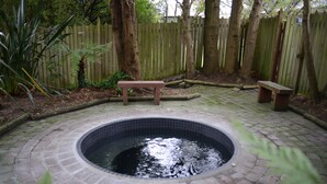 Una bañera de hidromasaje circular rodeada por un patio de ladrillo, con bancos de madera y una valla de madera al fondo.