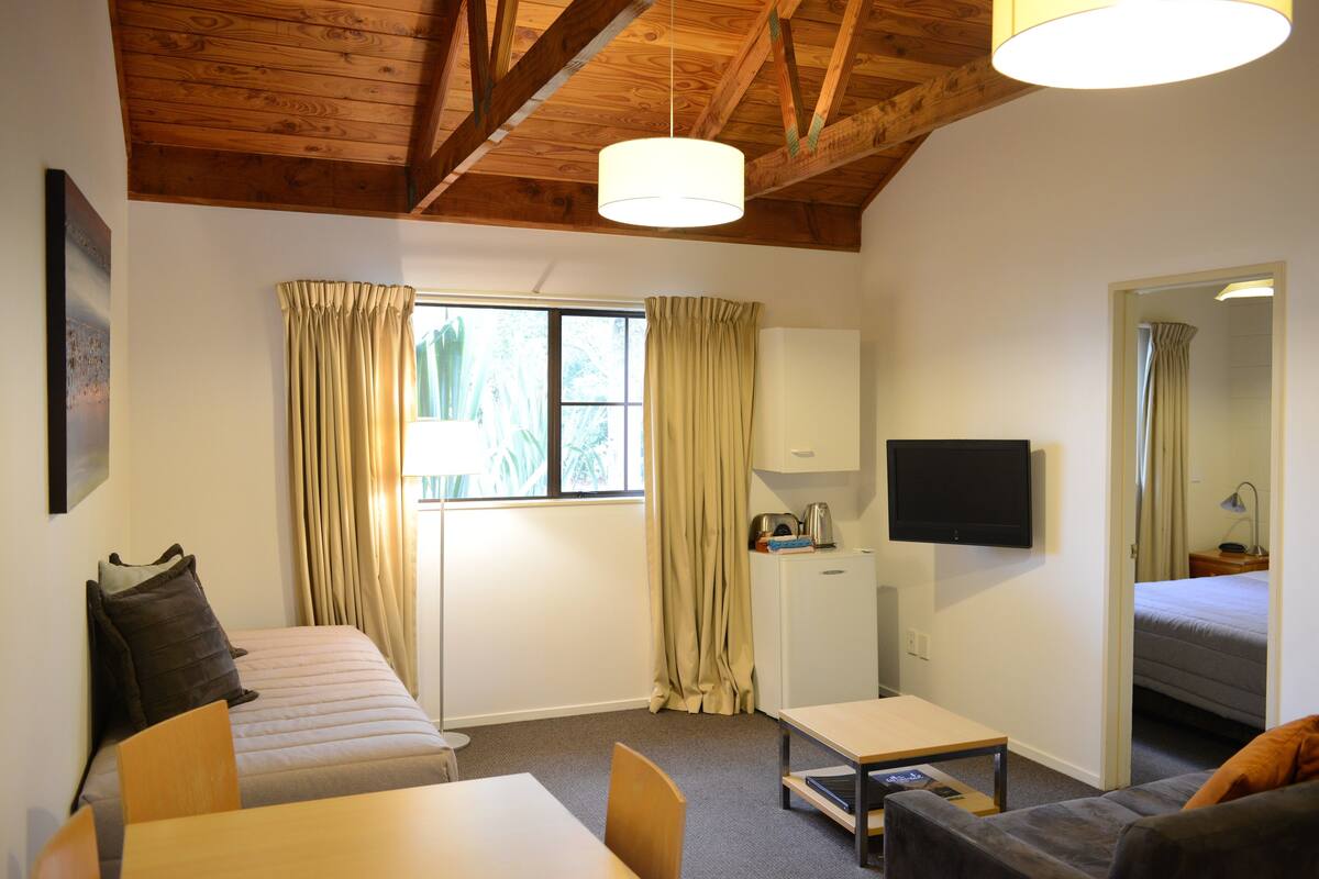 Una habitación con cama, sofá, mesa, televisor y ventana con cortinas.