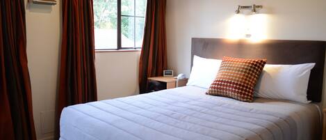 Una cama bien hecha con cabecera, una mesita de noche con lámpara y una ventana con cortinas.
