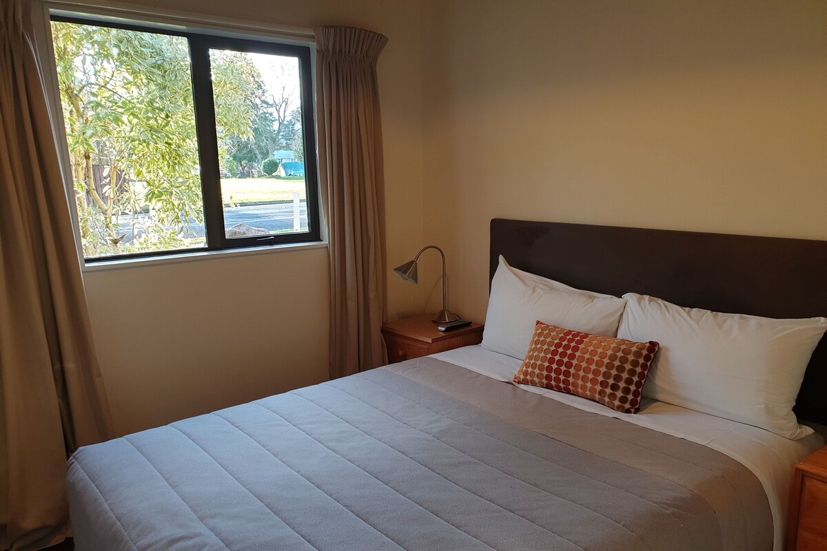 A hotel room with a large bed, a nightstand, a lamp, and a window with curtains.