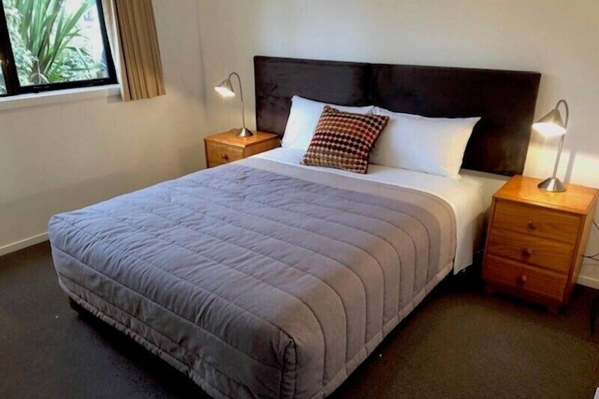 Habitación de hotel con una cama grande, dos mesitas de noche, un ventanal con cortinas y vistas a la vegetación.