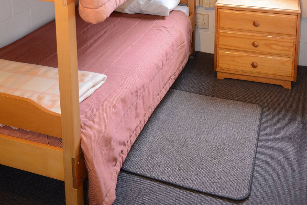 Una cama individual con ropa de cama rosa, una mesita de noche de madera con cajones y una ventana con cortinas.