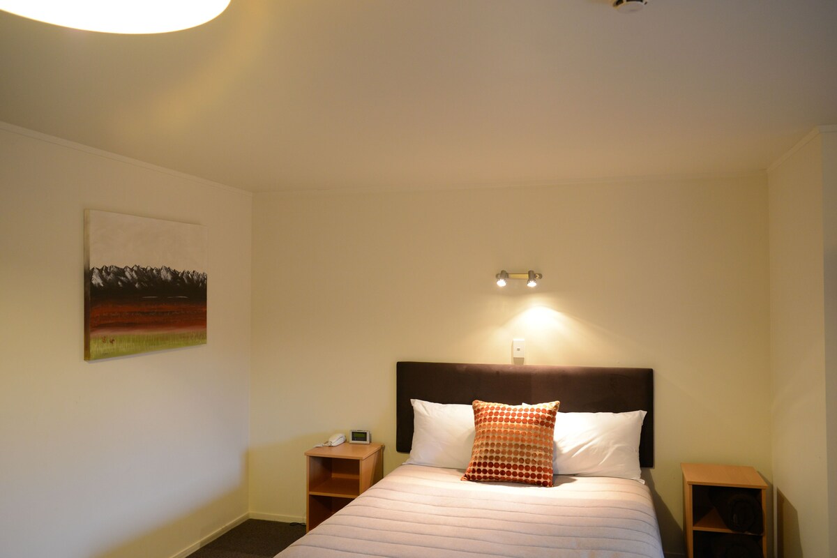 A hotel room with a bed, a nightstand, a wall-mounted light, and a painting.