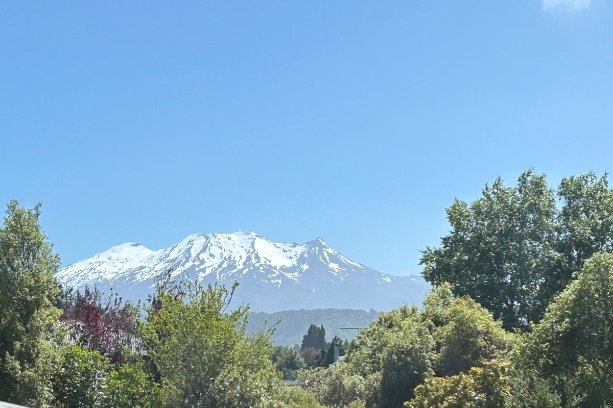 A snow-capped mountain with a clear sky, surrounded by dense green foliage.