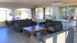 A spacious living area with a wooden coffee table, grey sofas, and a dining table with chairs. Large windows provide ample natural light and views of the outdoors. - Toukley Motor Inn (Toukley)