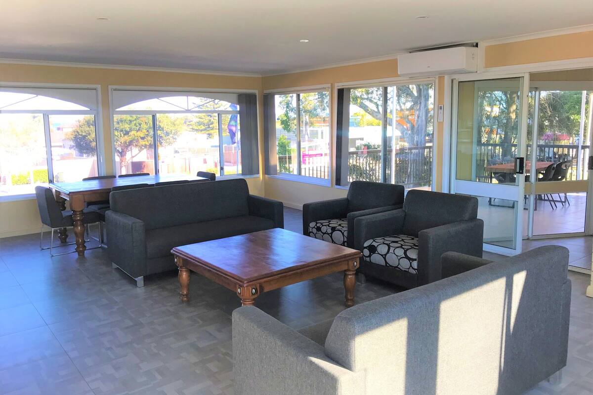 A spacious living area with a wooden coffee table, grey sofas, and a dining table with chairs. Large windows provide ample natural light and views of the outdoors.