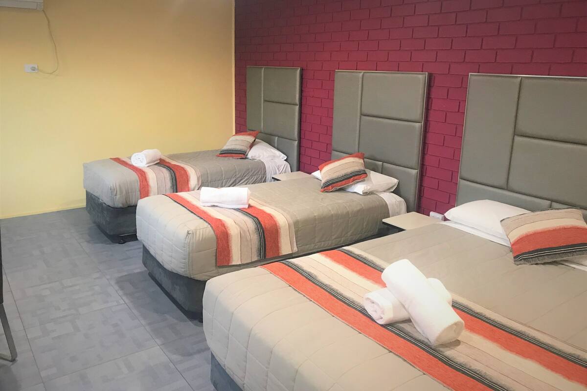 A hotel room with three beds, each with a headboard and pillows, and a red brick wall.