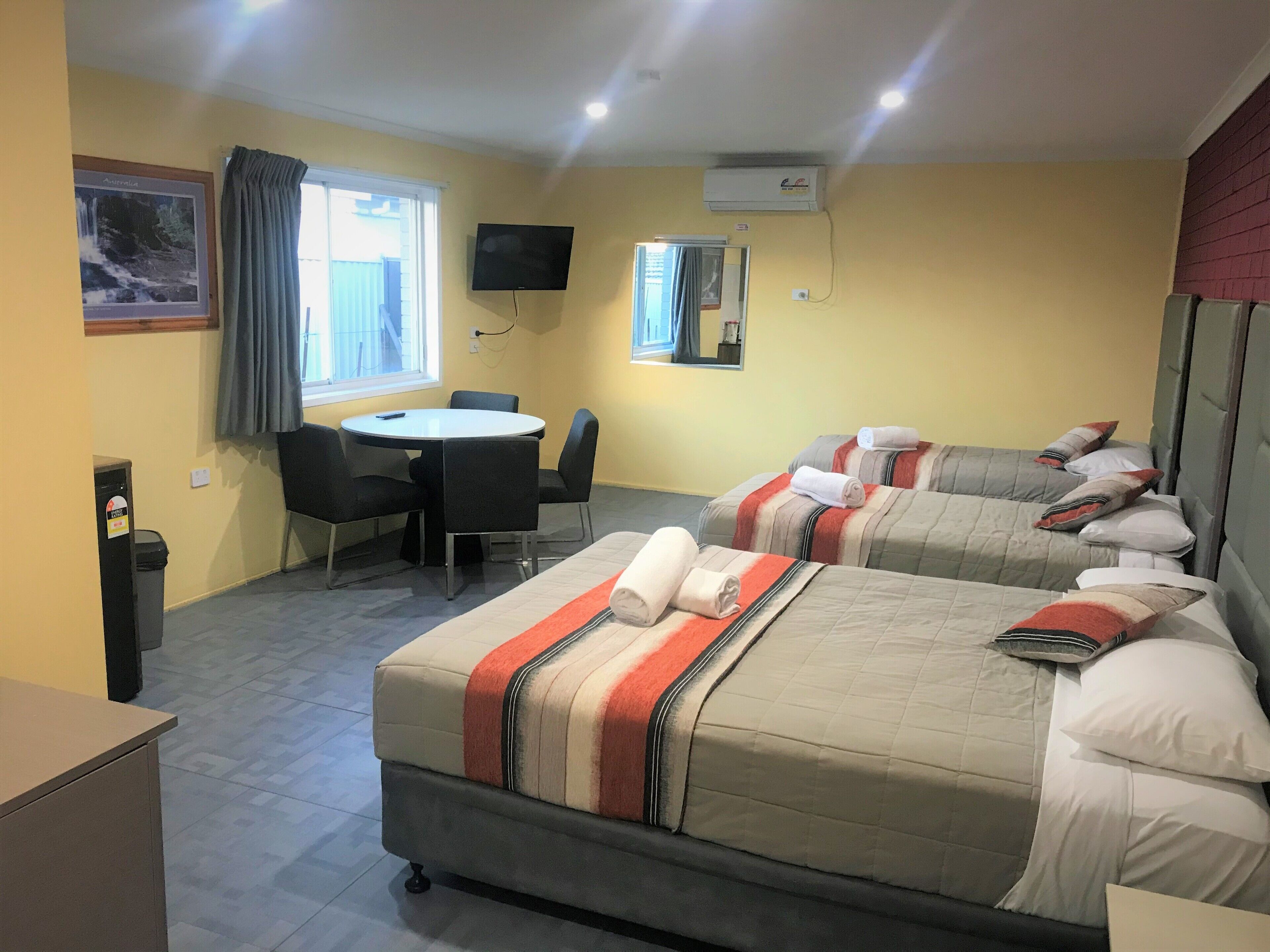 Habitación de hotel con dos camas, una mesita redonda con sillas, un televisor montado en la pared y una ventana con cortinas.
