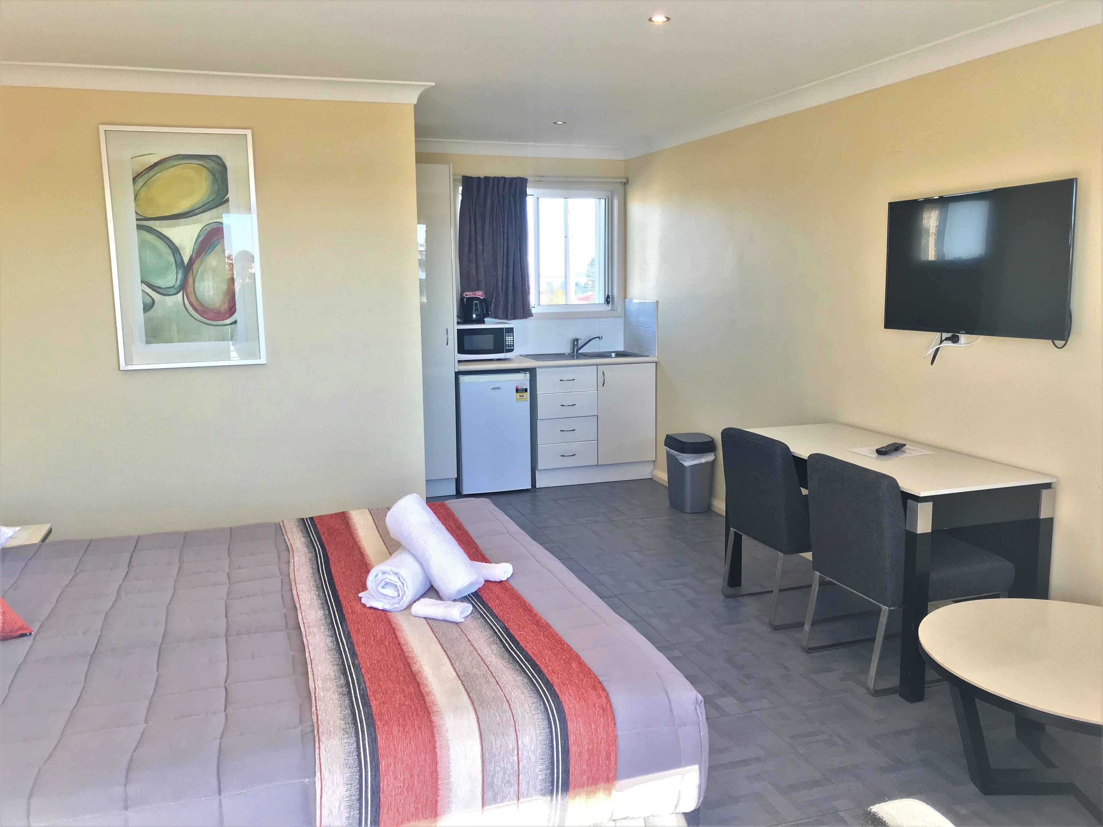 A hotel room with a bed, a desk, a chair, a TV, a kitchen area, and a painting on the wall.