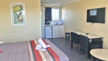 A hotel room with a bed, a desk, a chair, a TV, a kitchen area, and a painting on the wall.