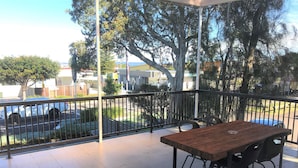 A covered outdoor area with a wooden table and chairs, a metal railing, and a view of a street with parked cars and trees. - Toukley Motor Inn (Toukley)