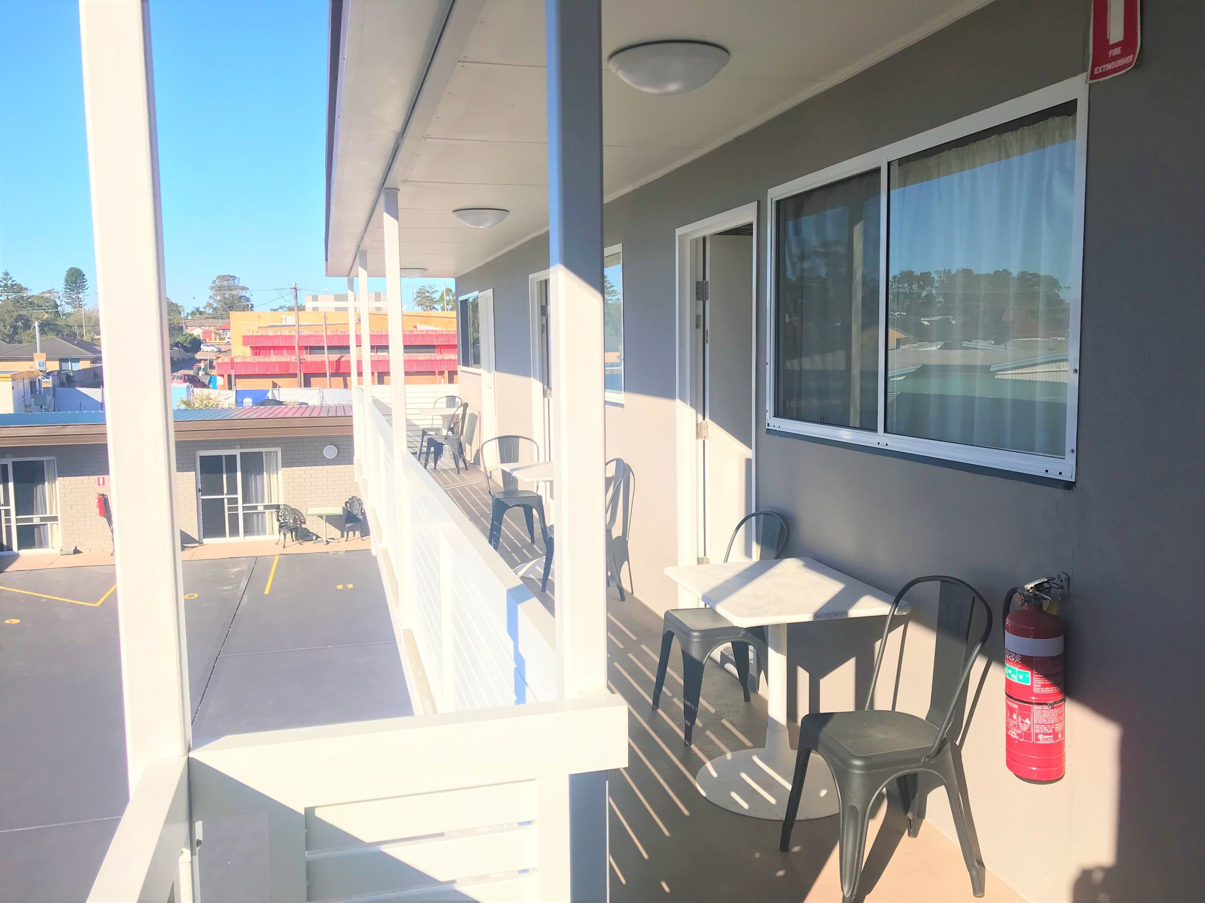 Un balcón con mesa y sillas, un extintor y vistas a un estacionamiento y edificios.