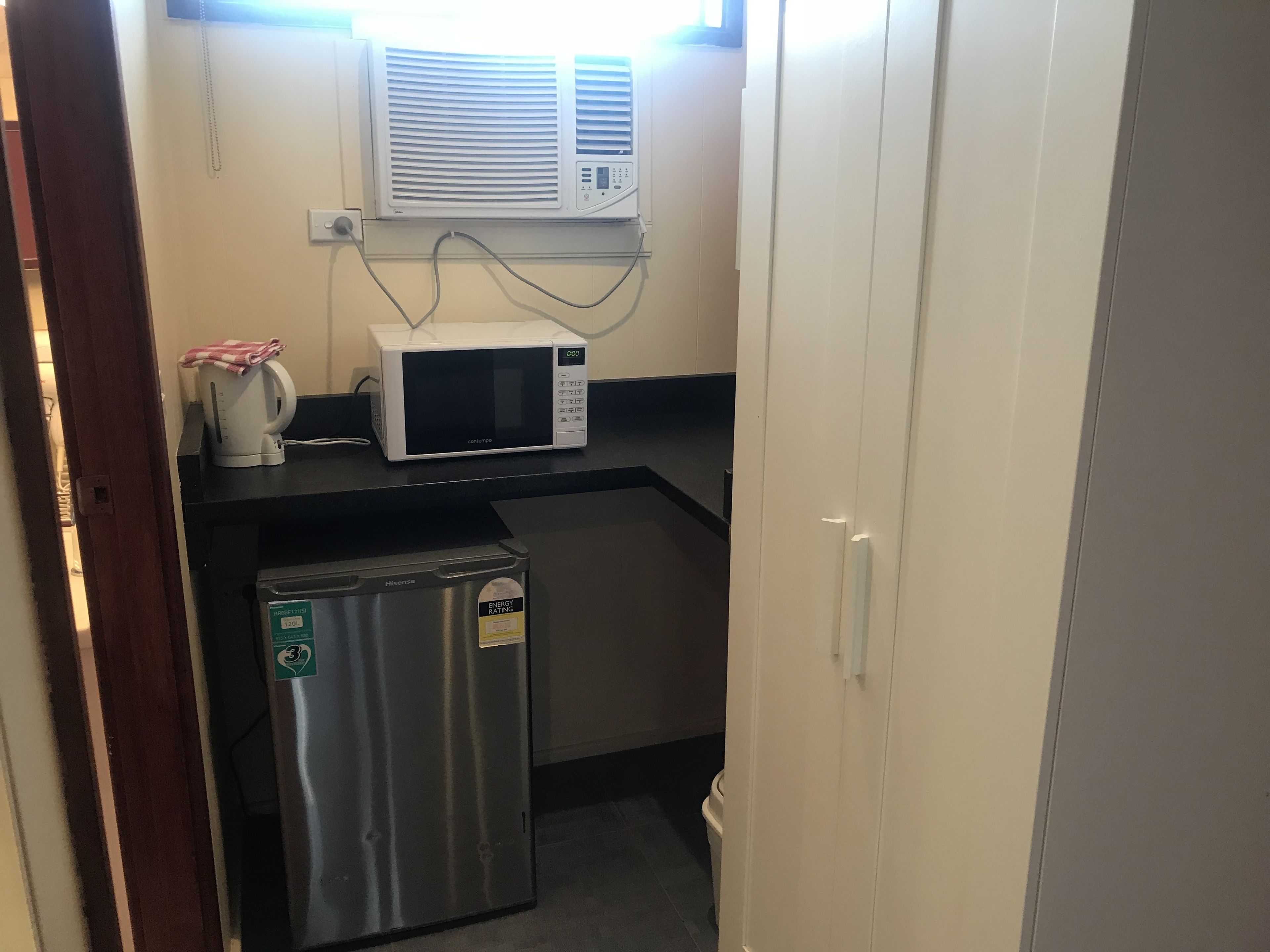 A compact kitchenette with a microwave, a small refrigerator, and a window-mounted air conditioner.