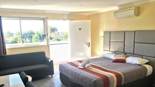 A hotel room with a bed, a sofa, a table, and a window with a view of greenery. - Toukley Motor Inn (Toukley)