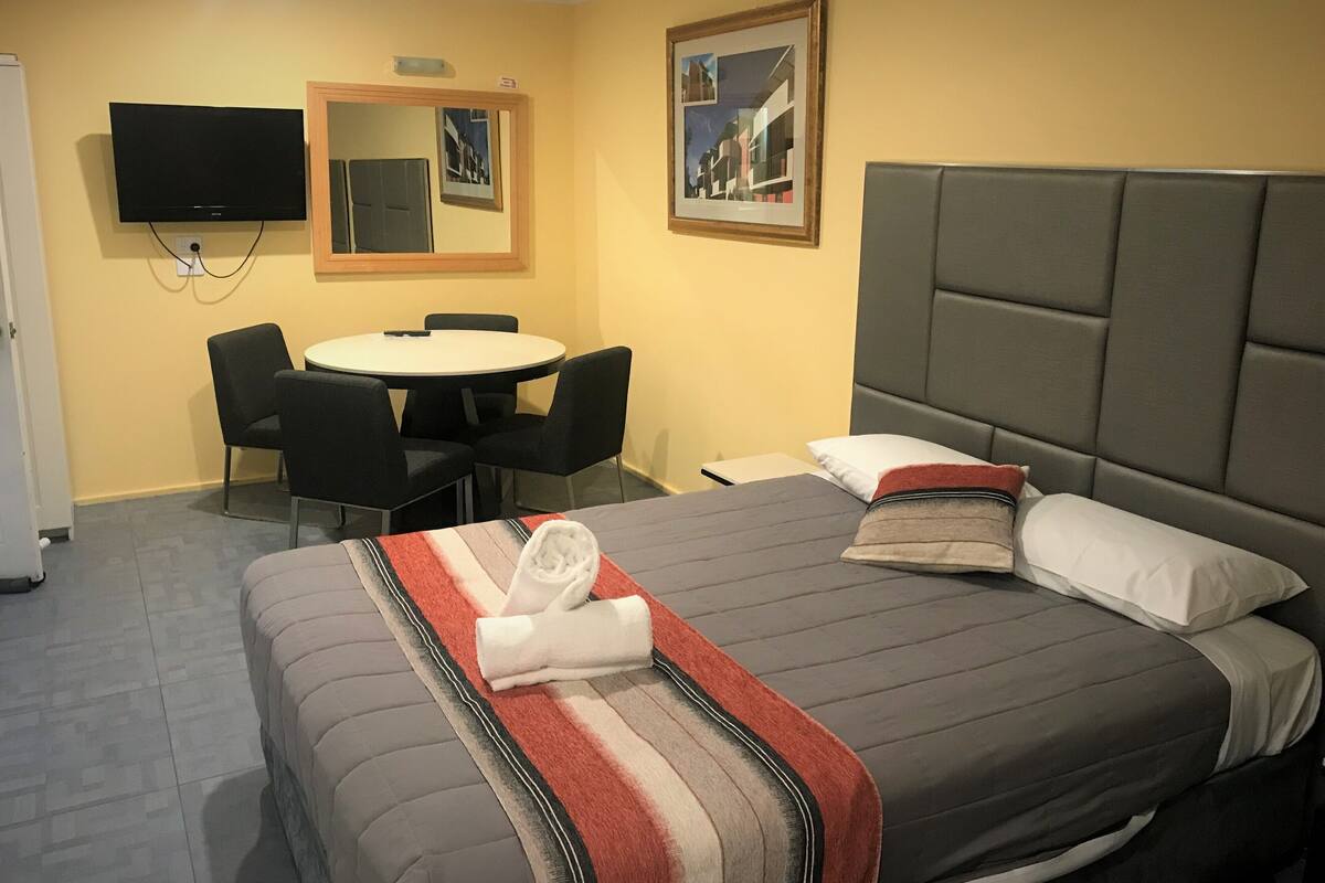 A hotel room with a bed, a round dining table with chairs, a television mounted on the wall, and a framed picture above the bed.