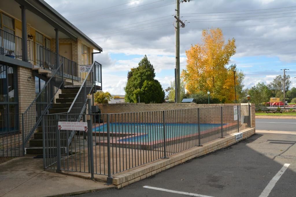 A motel with a fenced pool area, a staircase leading to the entrance, and a large tree with autumn leaves in the background.