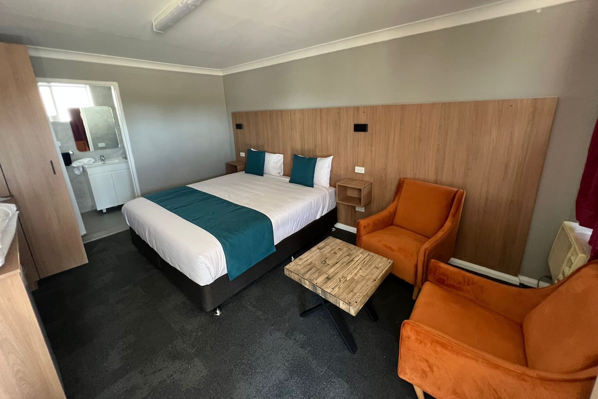 Une chambre d’hôtel avec un lit, une table de chevet en bois, un fauteuil orange, une petite table en bois et un miroir.