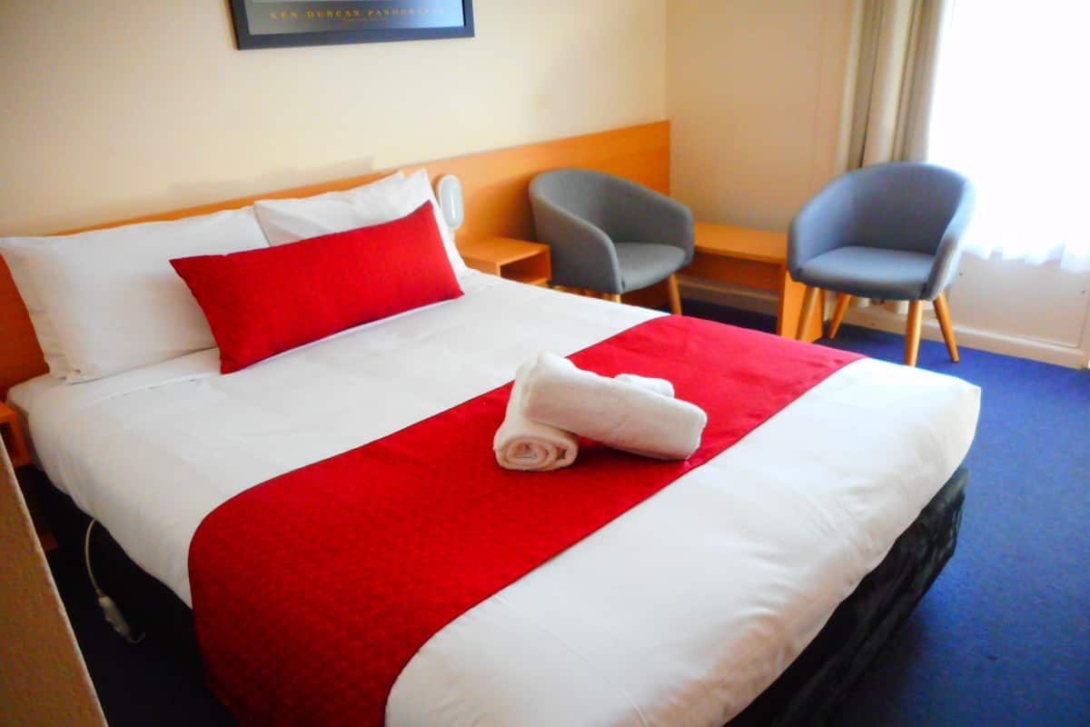 Une chambre d’hôtel avec un lit bien fait, deux fauteuils, une petite table et une fenêtre avec des rideaux.