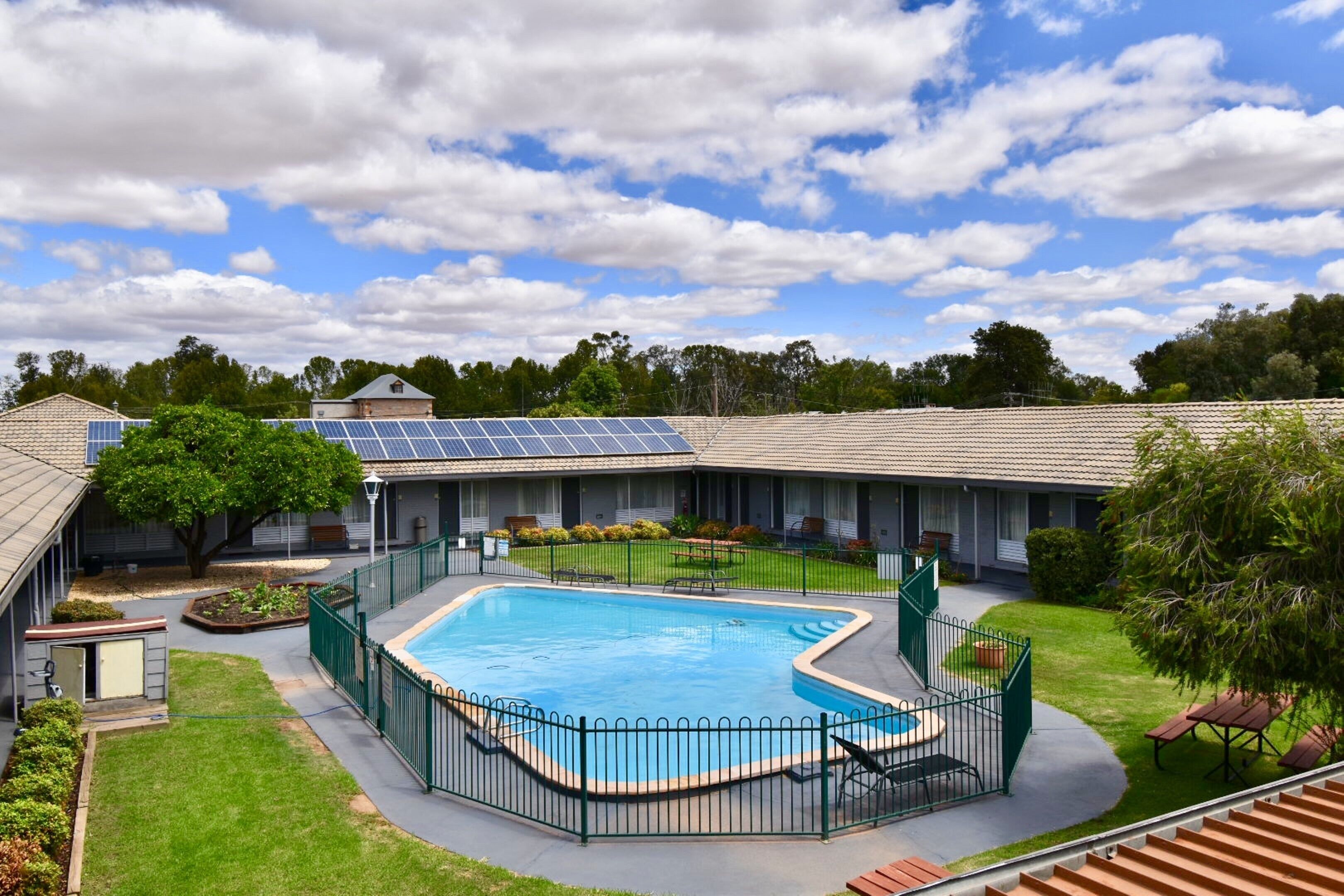 A swimming pool surrounded by a green fence, with a building in the background and a clear sky.