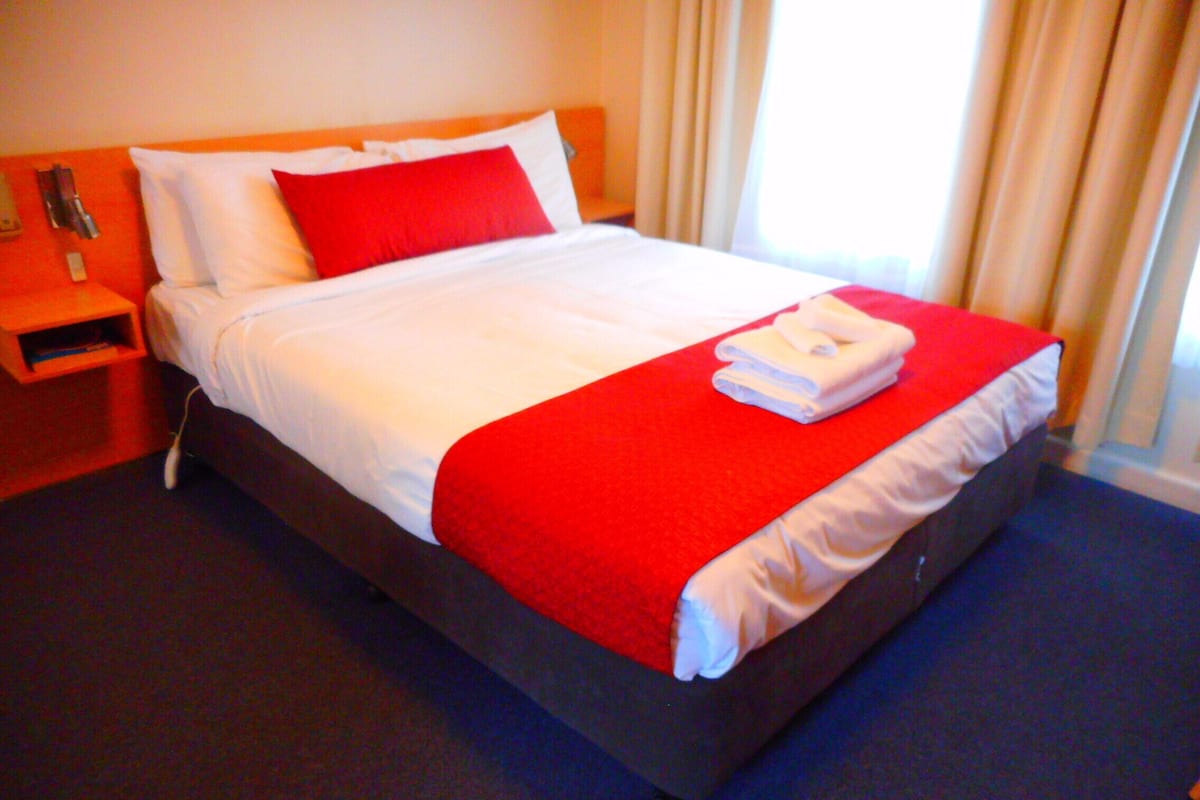Un lit bien fait, avec une couverture rouge et blanche, une serviette blanche pliée et un oreiller rouge.