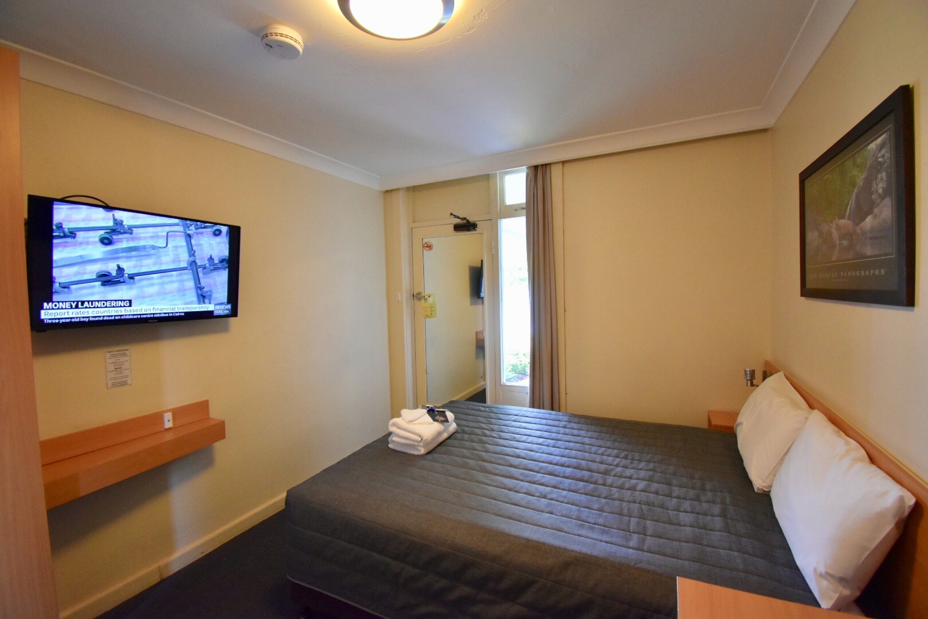 Hotellihuone, jossa on sänky, seinälle asennettu televisio, ovi, joka johtaa kylpyhuoneeseen, ja seinällä oleva kehystetty kuva.