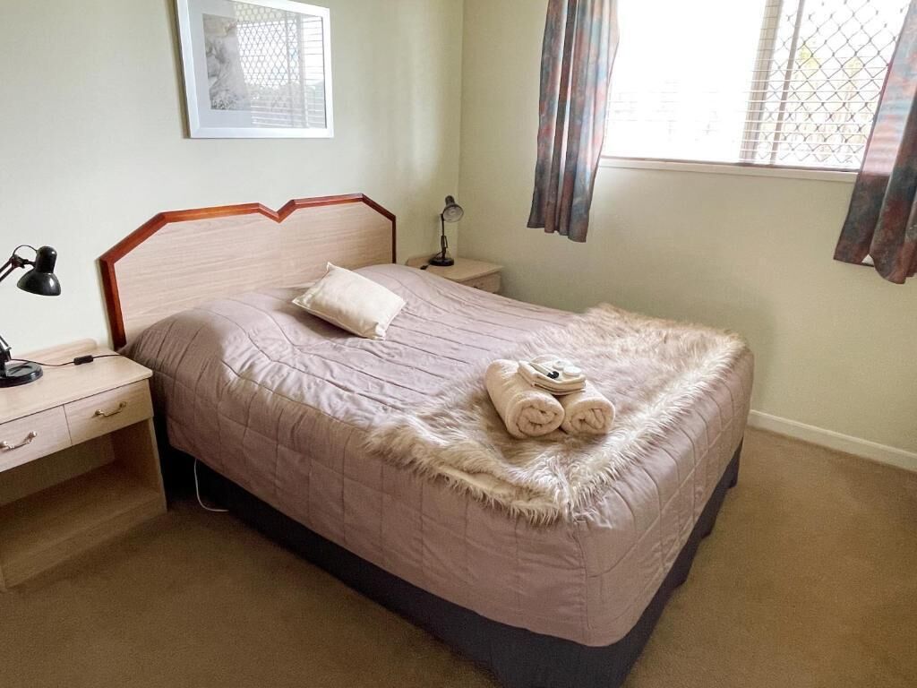 A neatly made bed with a fluffy blanket and rolled towels, a bedside lamp, a small table with a lamp, and a framed picture on the wall.