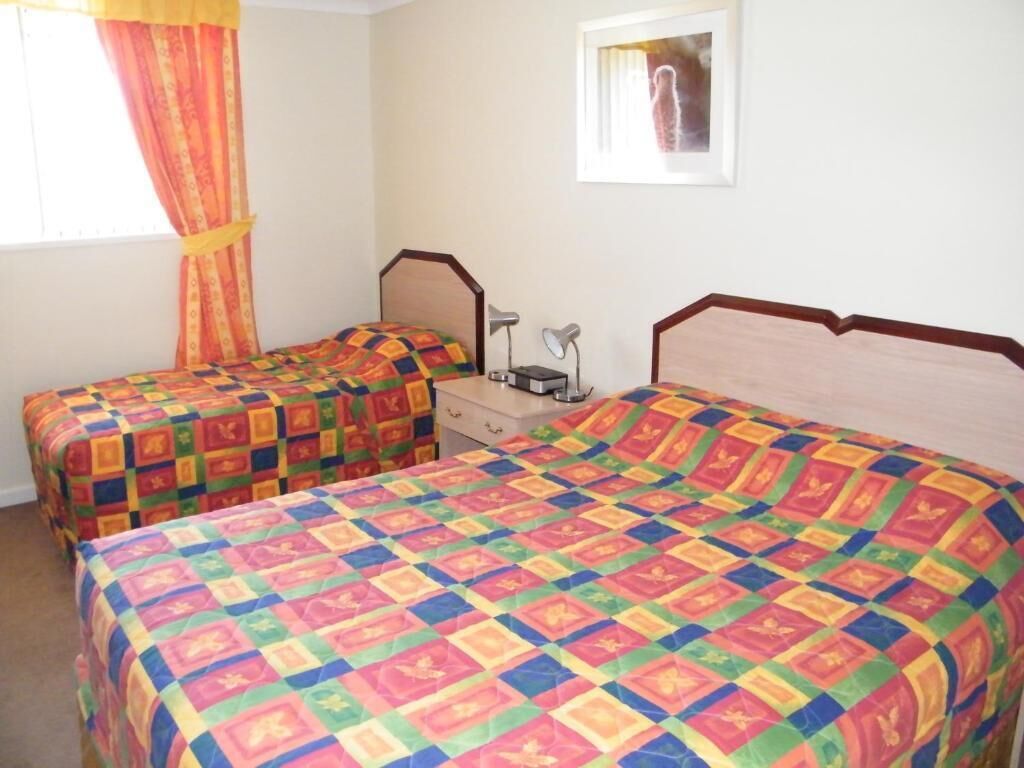 Una habitación con dos camas individuales, cada una con una colcha de colores, una mesita de noche con una lámpara y un cuadro en la pared.