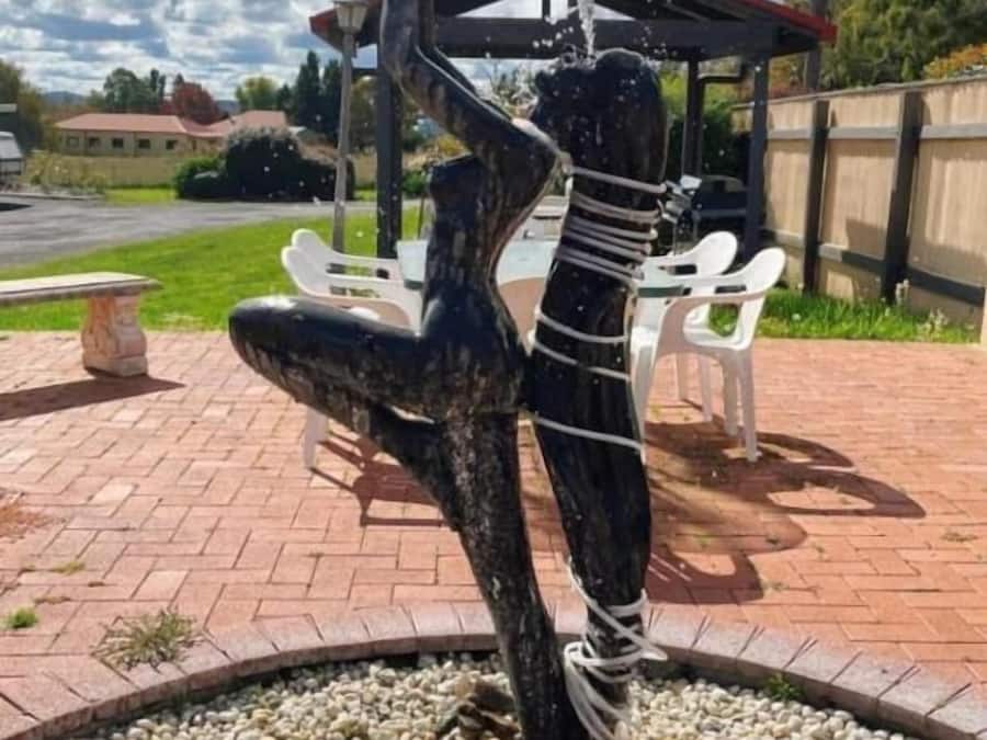 Eine Skulptur einer Figur, die eine Tasse hält, umgeben von einem kreisförmigen Steinrondell, mit einem überdachten Sitzbereich im Hintergrund.