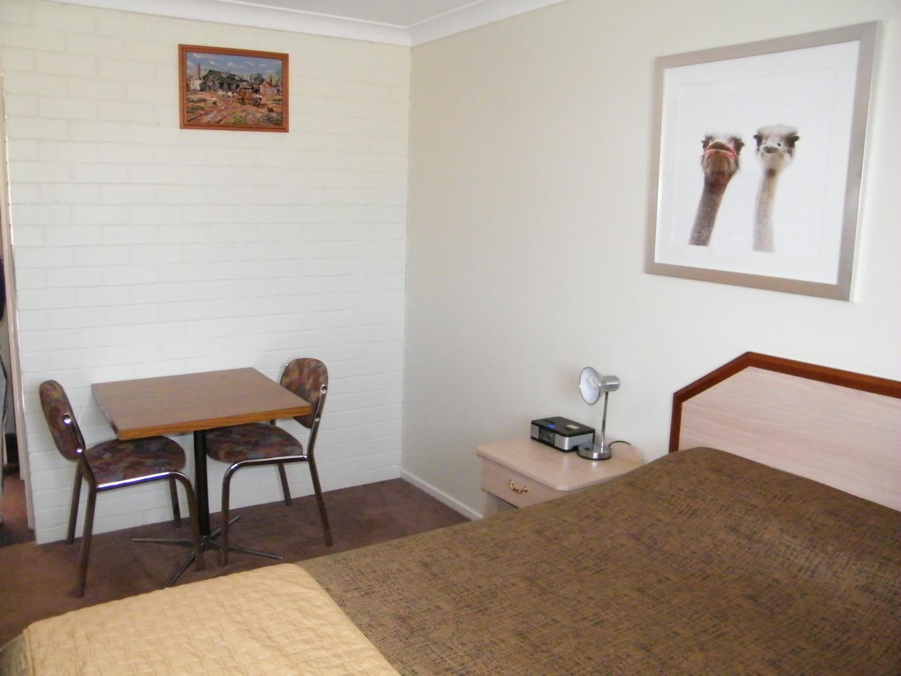 A room with a bed, a desk, two chairs, a nightstand with a lamp, and a framed picture on the wall.