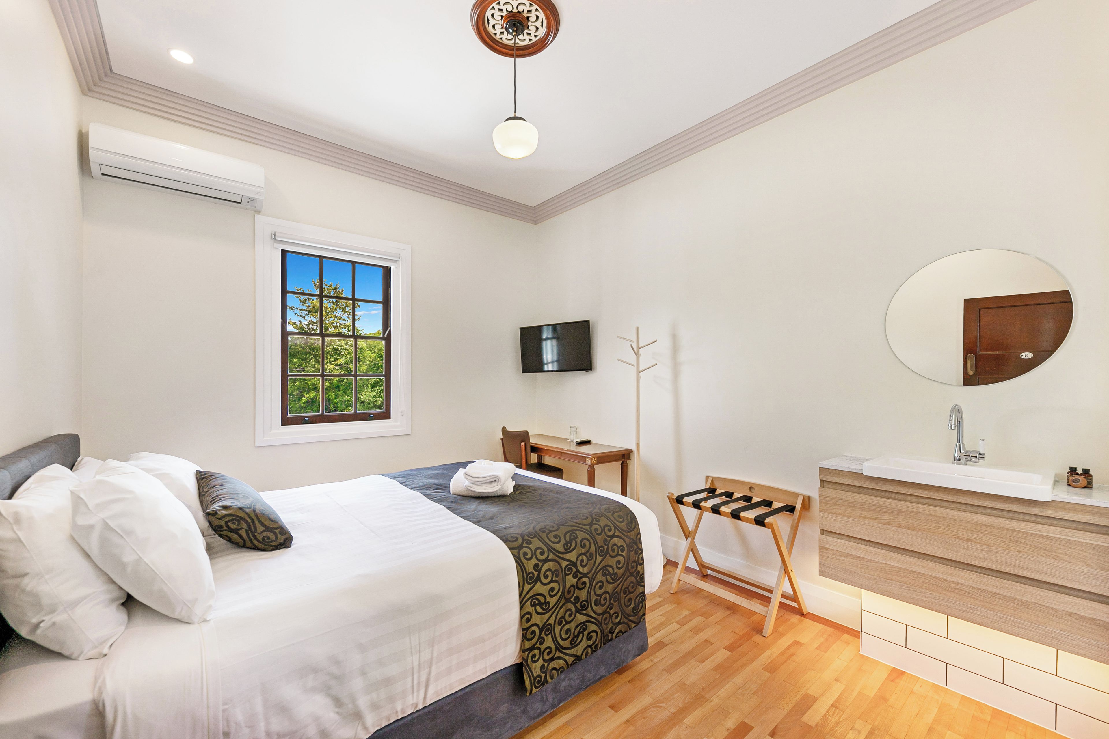 Habitación de hotel con cama, escritorio, televisión, espejo y baño con lavabo y tocador de madera.